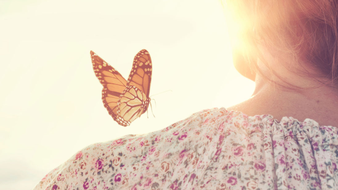 Close-up rear view of a butterfly landing on a woman's shoulder with glowy sunlight in the background.